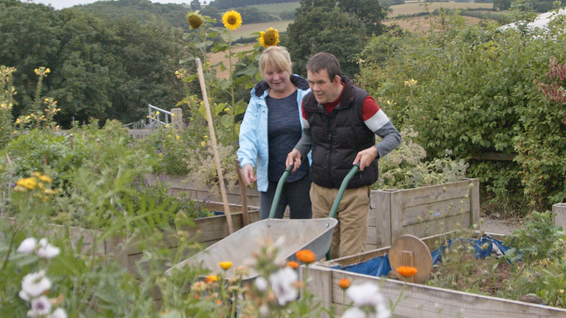 Pretty garden setting with flowers with two people at the centre, man pushes a wheelbarrow while woman looks on