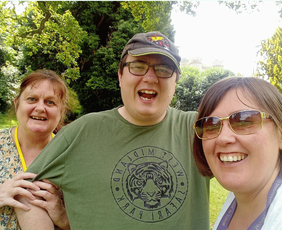 Three people smiling outdoors in front of trees and a building. The person in the middle wears a green t-shirt with a tiger design and a baseball cap. The person on the right wears sunglasses.