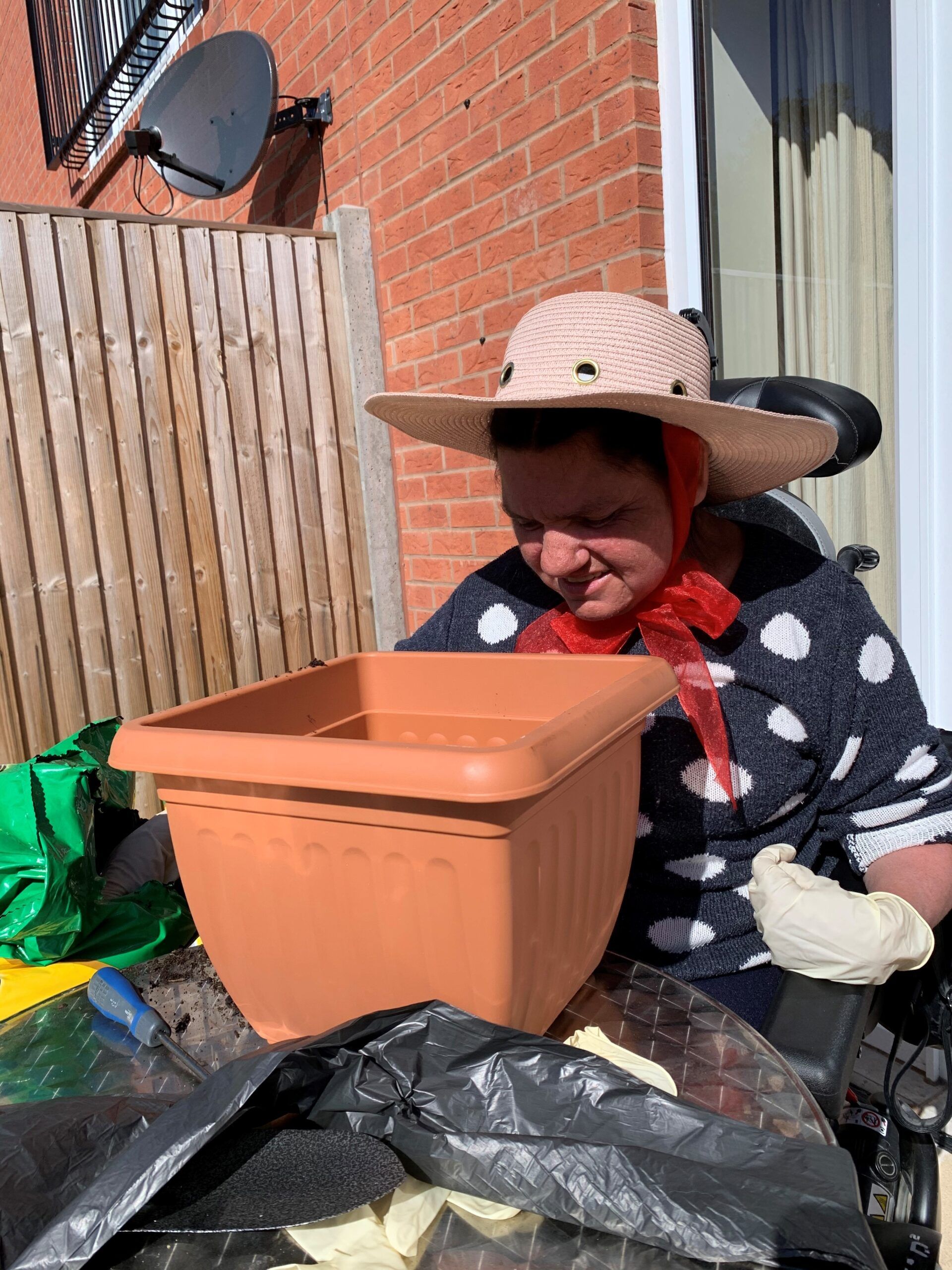 woman seated in a wheelchair sits at a table to do some gardening wearing a large hat to protect from the sun