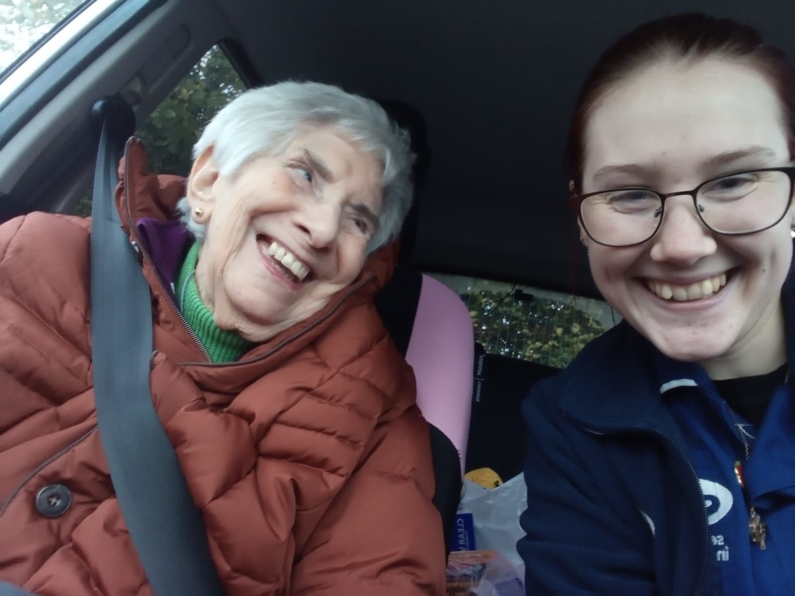 older woman in a car wearing a seat belt looks at her younger passenger both smiling