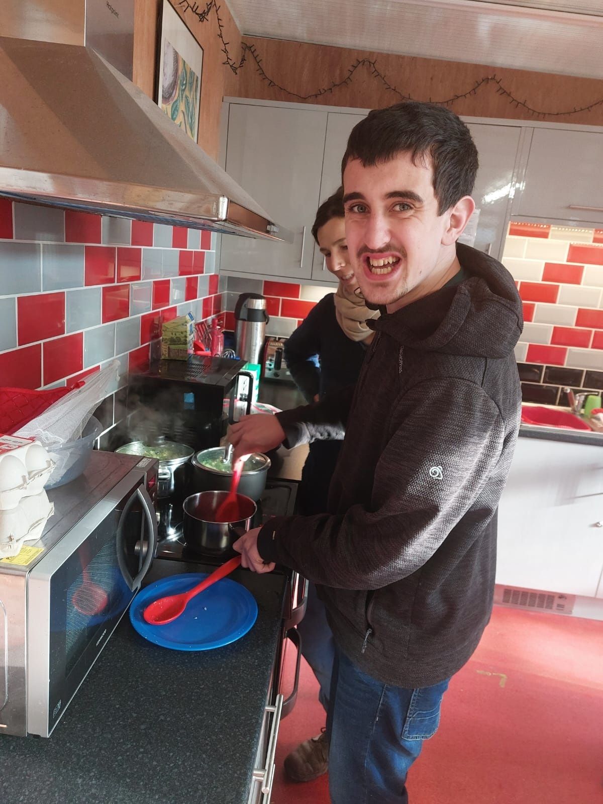 Young man stirs a saucepan with support worker looking on