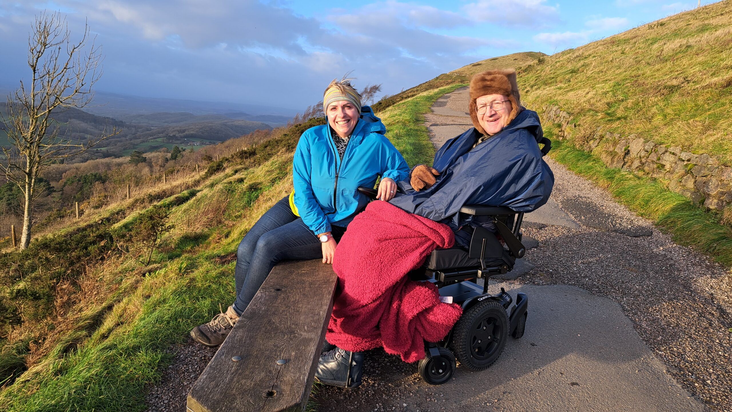 Picturesque setting on a hillside, distant views are enjoyed by a woman sitting on a bench and a man in a wheelchair.